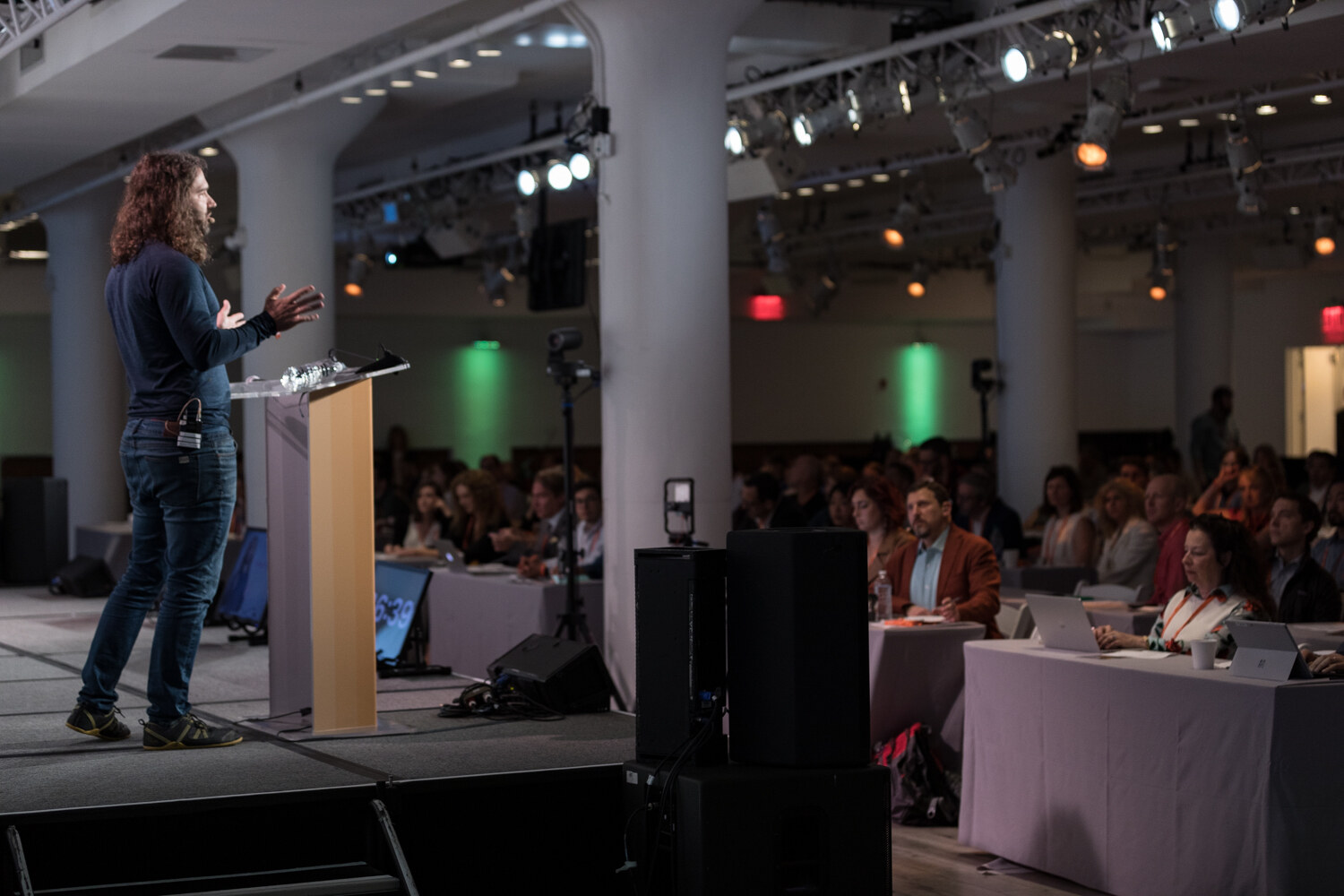 Wide view of the room with the man in a navy henley at the lectern and attendees at draped tables
