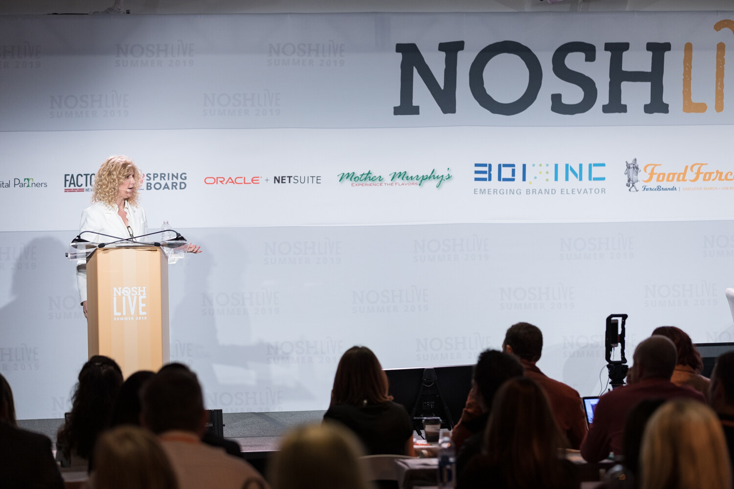 Woman in a white blazer at the NOSH LIVE lectern with audience heads in the foreground