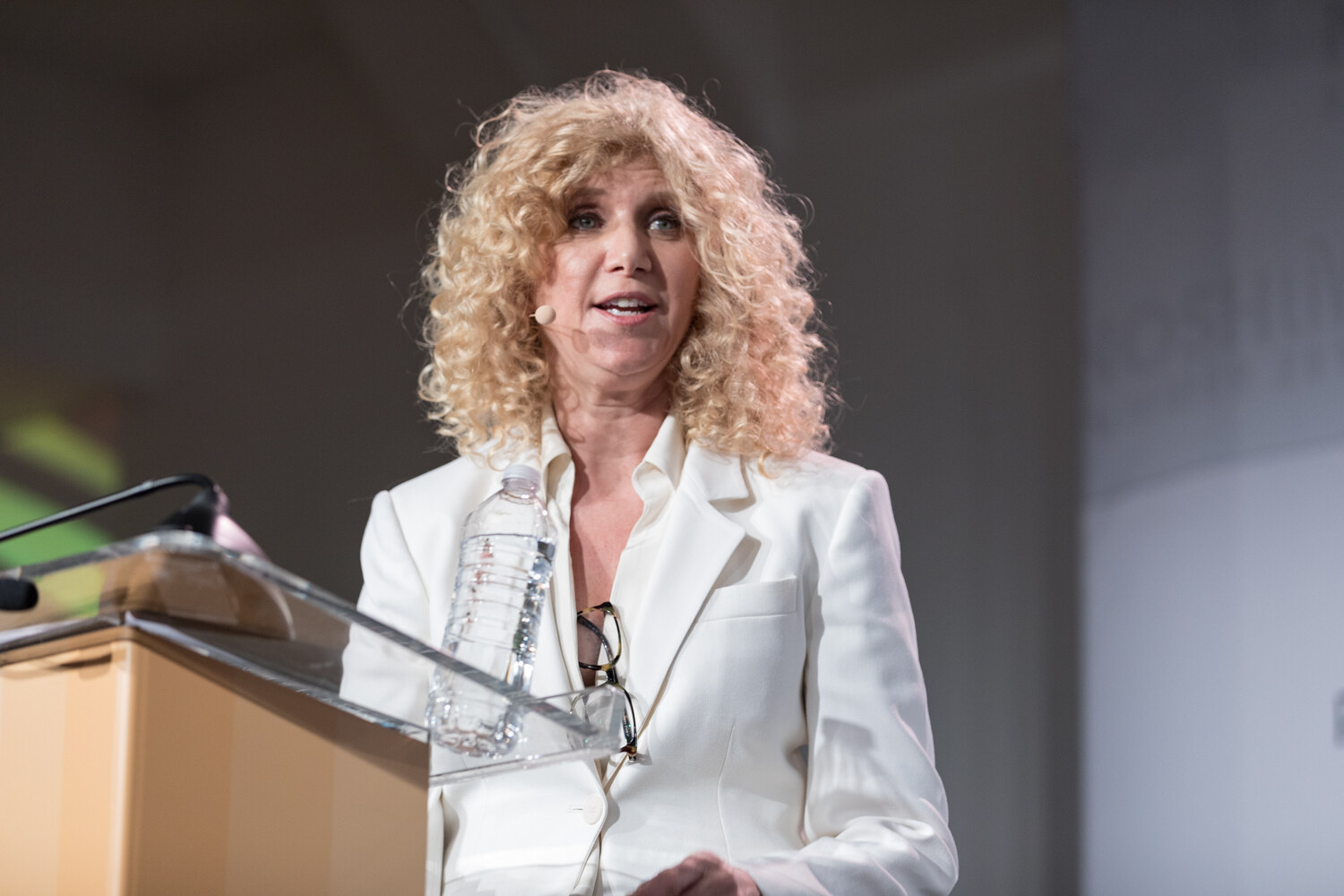 Woman with curly blond hair in a white blazer at the lectern beside a water bottle