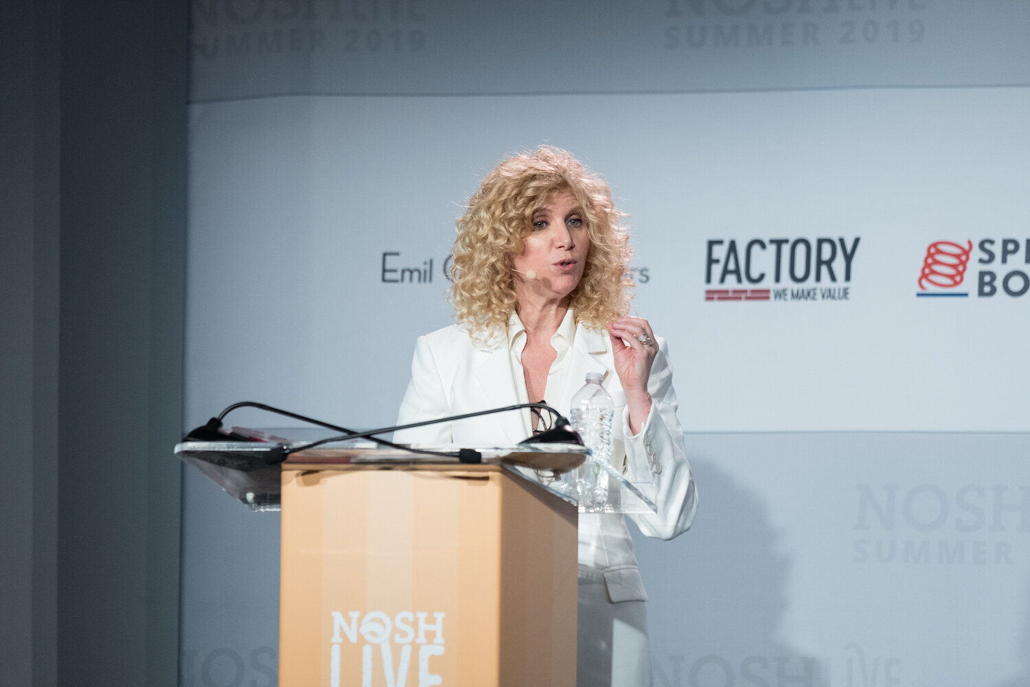 Woman with curly blond hair in a white blazer at the NOSH LIVE lectern in front of the Factory and Spring Board logos