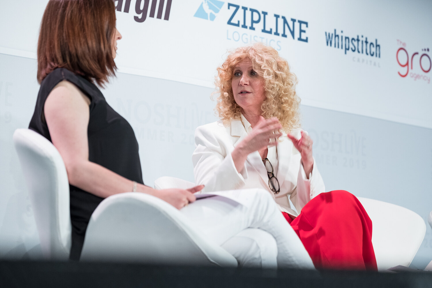 Woman in a white blazer and red pants gesturing with both hands as the interviewer in black listens