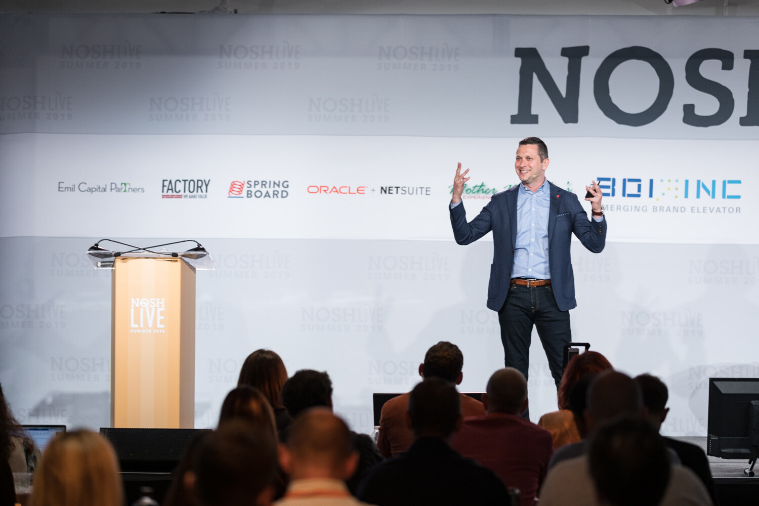 Speaker in a navy blazer and light blue shirt with open hands in front of the NOSHLIVE sponsor wall, audience heads in the foreground