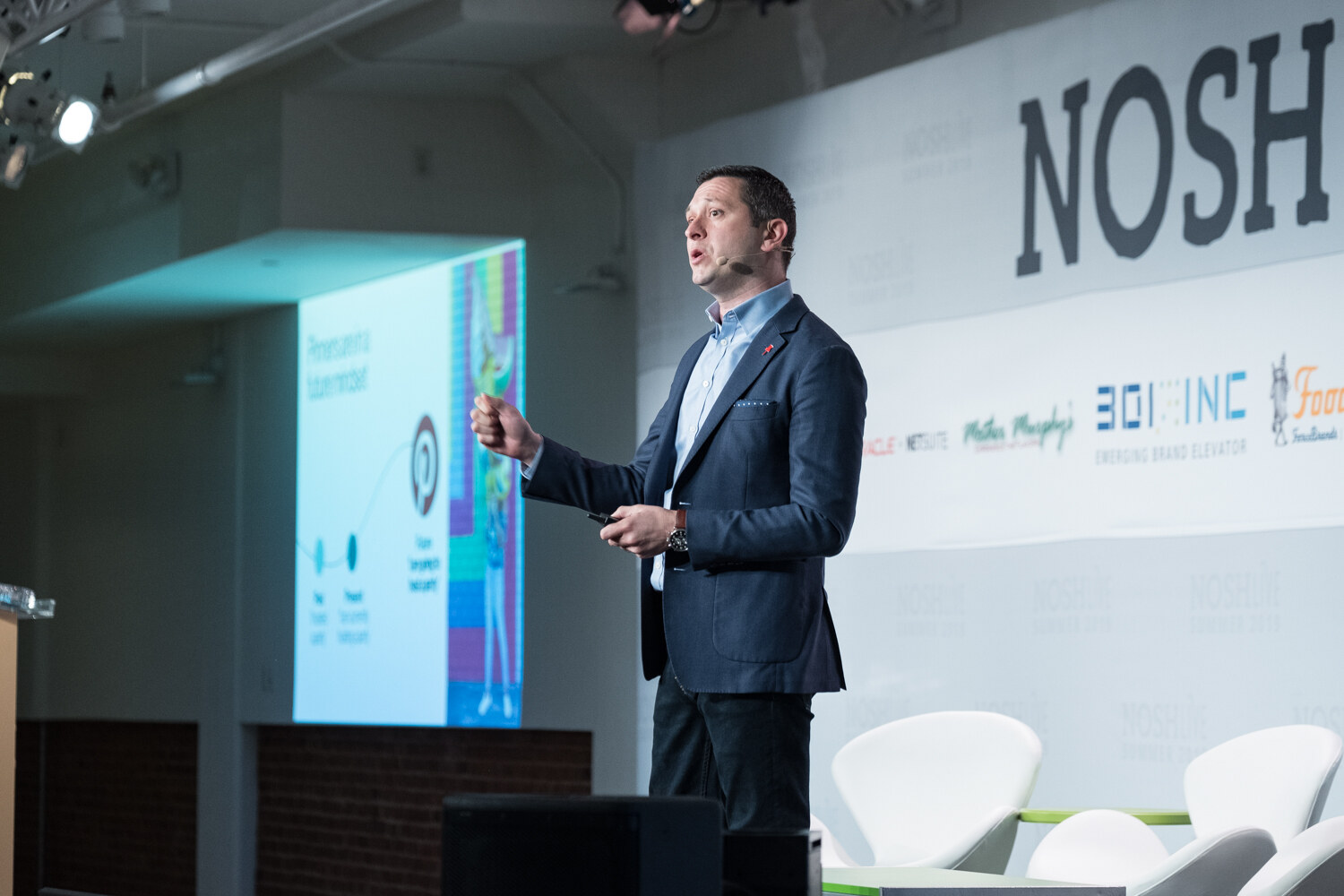 Speaker in a navy blazer gesturing beside a colorful projected slide on stage