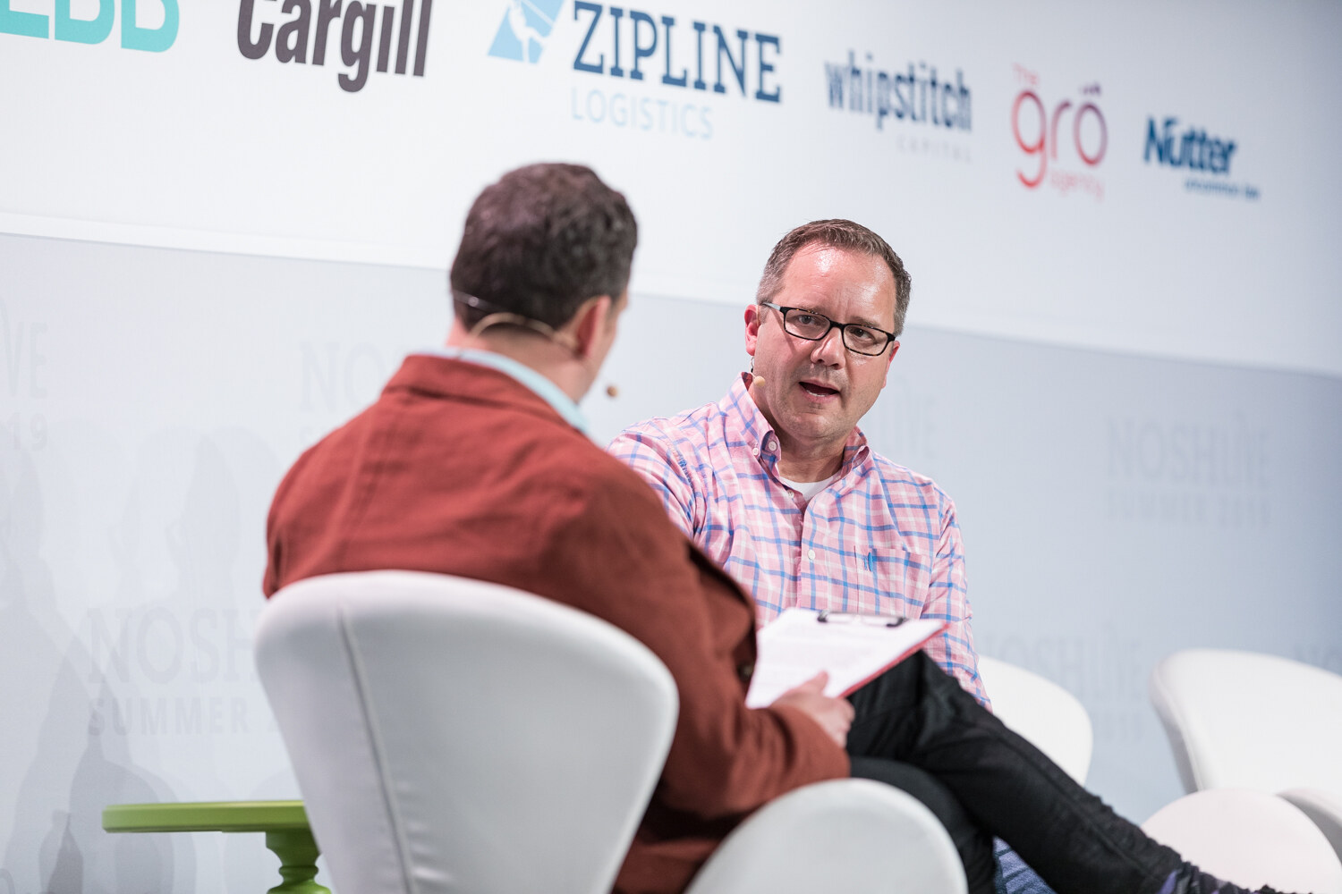 Host in a rust jacket seen from behind and the speaker in a pink plaid shirt seated on stage in conversation