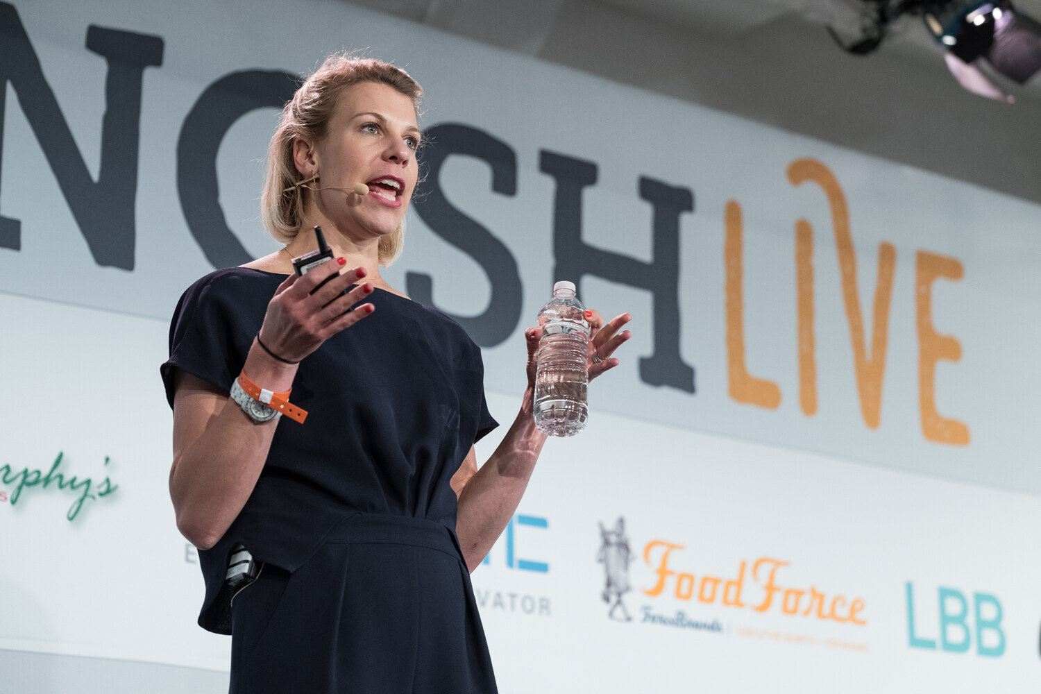 Woman in a dark jumpsuit gesturing with a water bottle in front of the NOSHLIVE wall