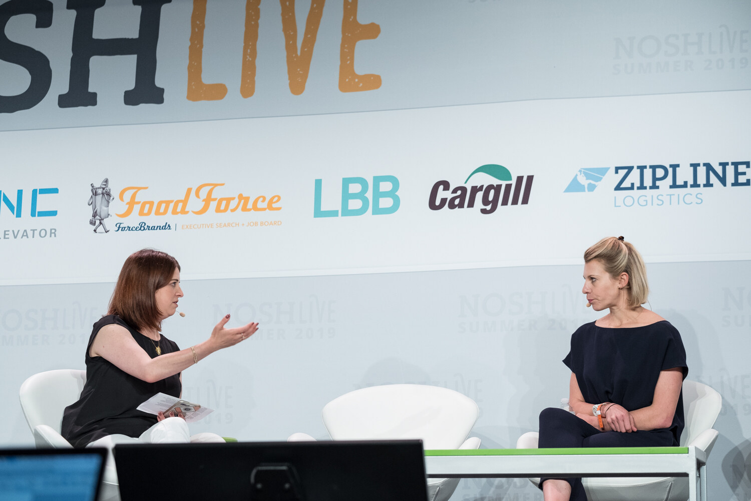 Moderator in black gesturing toward a woman in a dark jumpsuit seated in white swan chairs on the NOSHLIVE stage