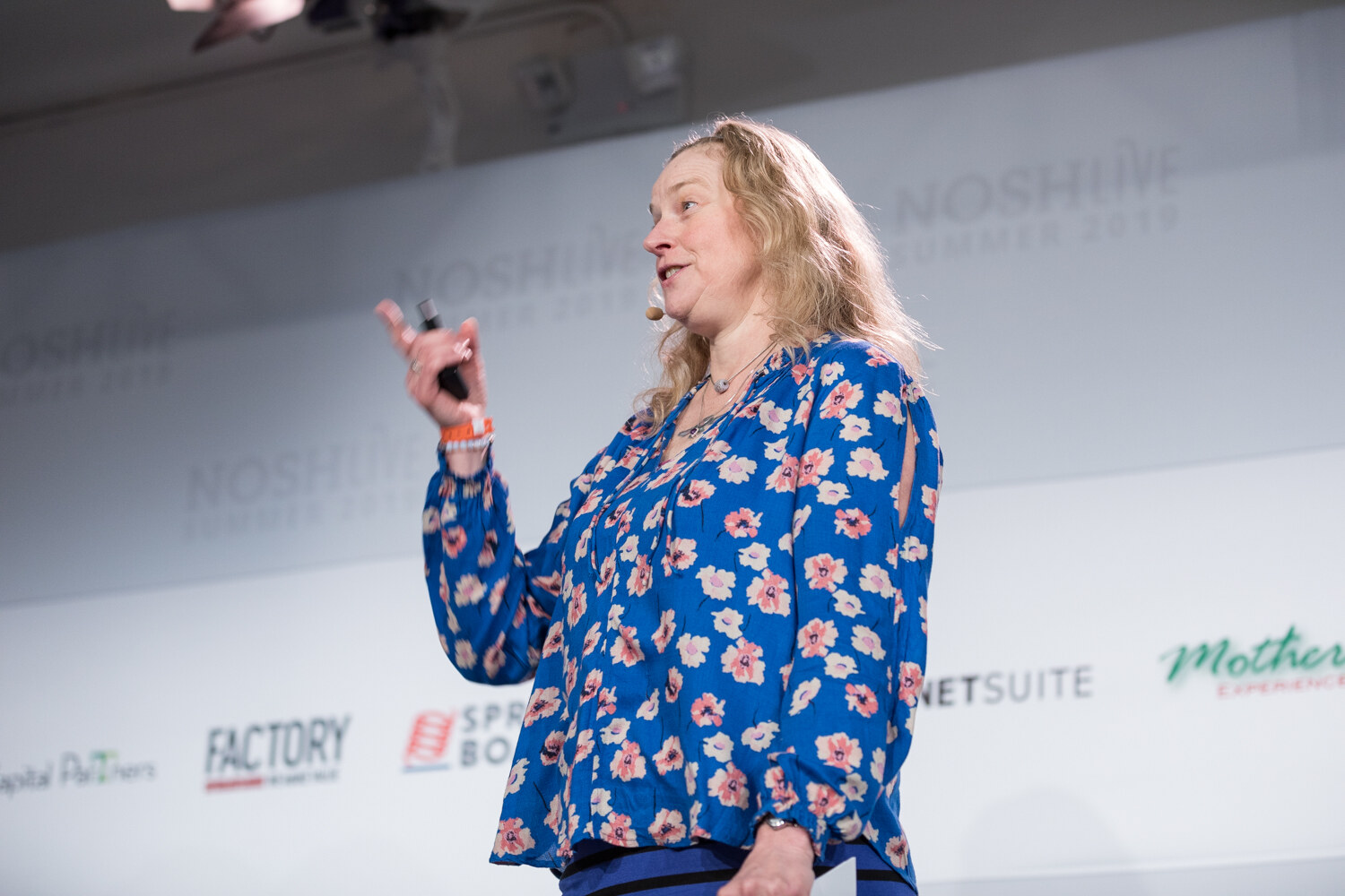 Woman in a blue floral blouse holding a clicker and gesturing in front of the sponsor wall