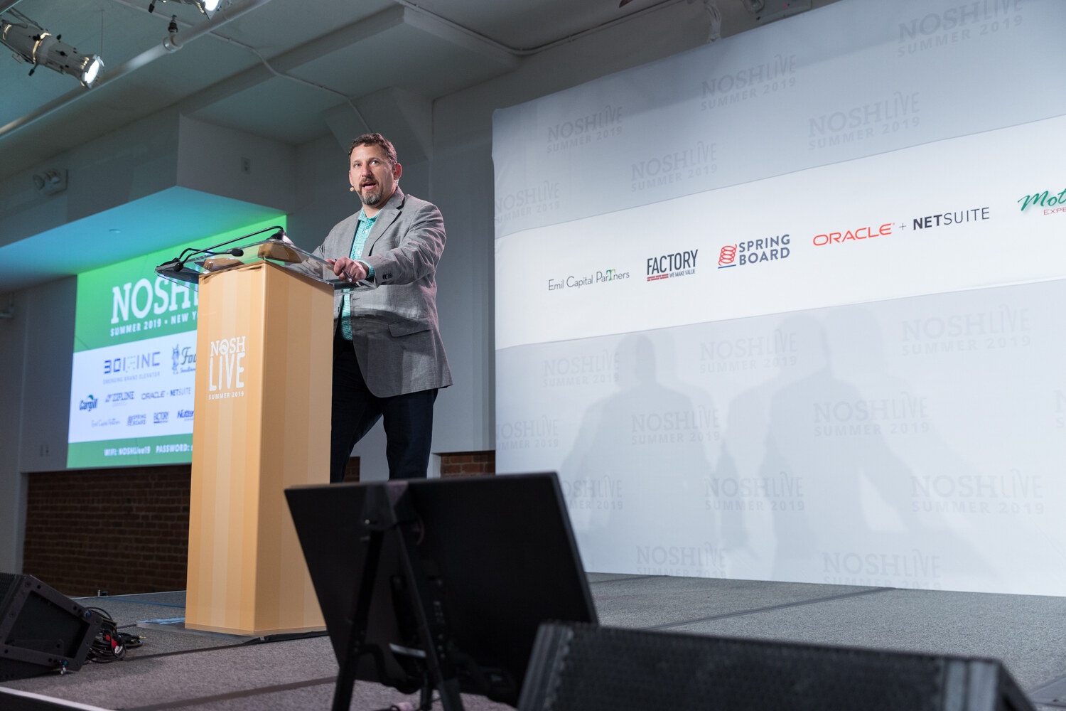 Bearded host in a gray blazer and teal shirt at the NOSH LIVE lectern in front of a green NOSH LIVE New York screen