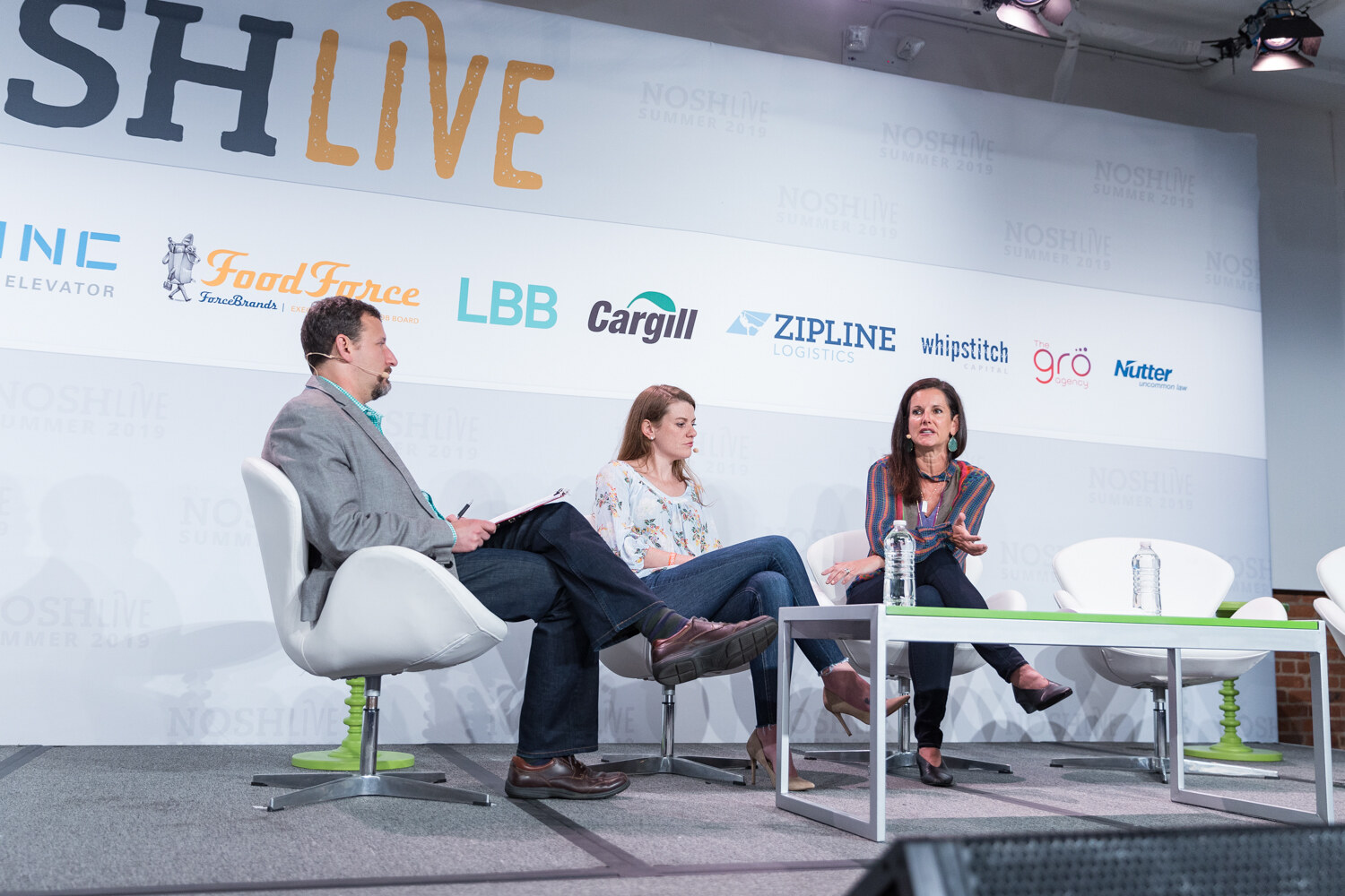 Moderator and two women seated in white swan chairs on stage in front of the NOSHLIVE wall