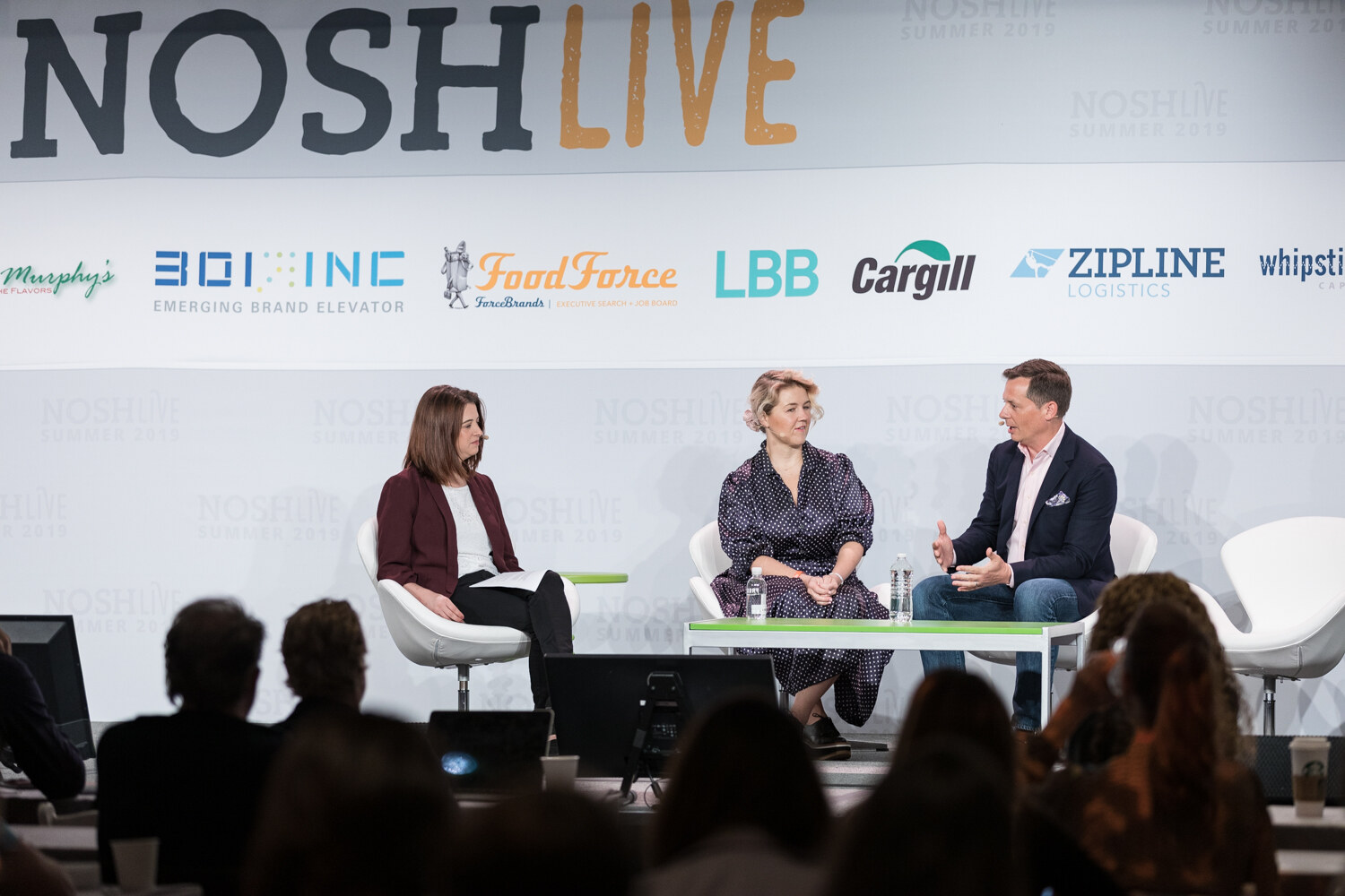Wide view of the Founders and CEOs panel with the moderator, a woman in a polka-dot dress and a man in a navy blazer seated on stage, audience heads in the foreground