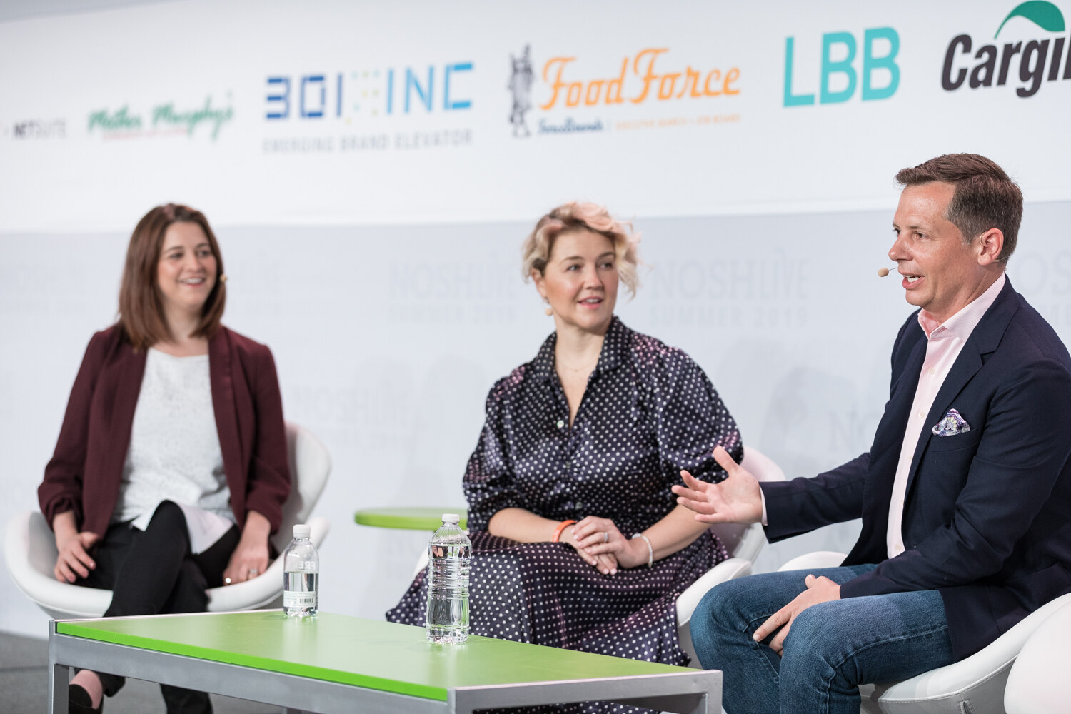 Man in a navy blazer gesturing toward the woman in a polka-dot dress while the moderator in burgundy listens