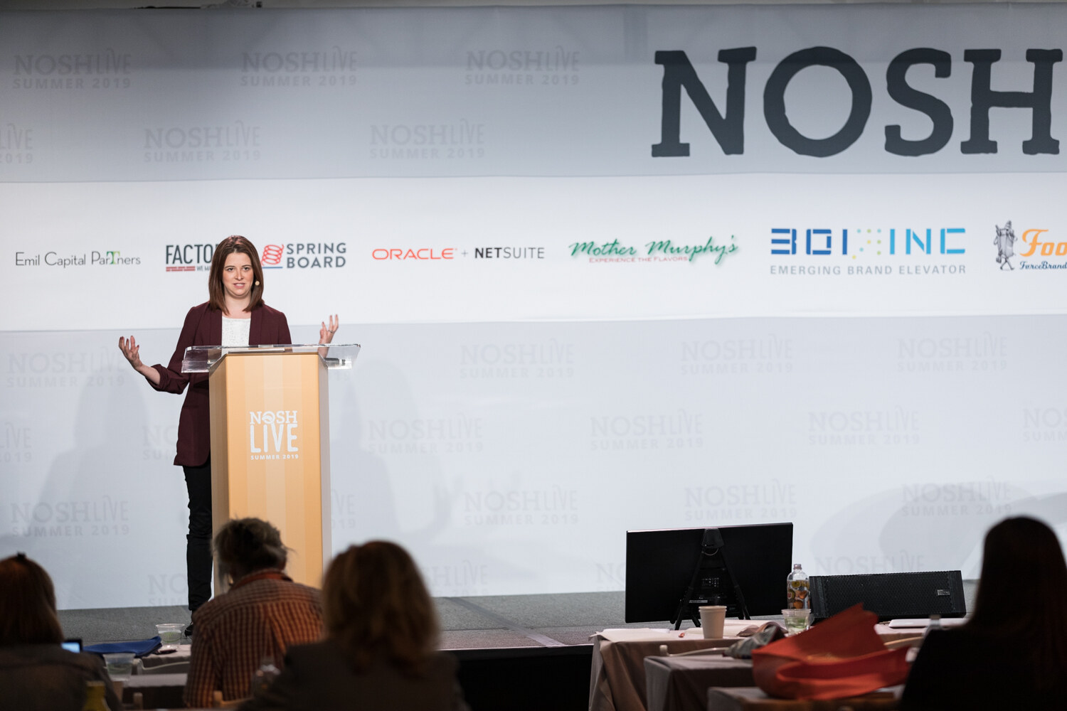Woman in a burgundy blazer at the NOSH LIVE lectern with both arms open