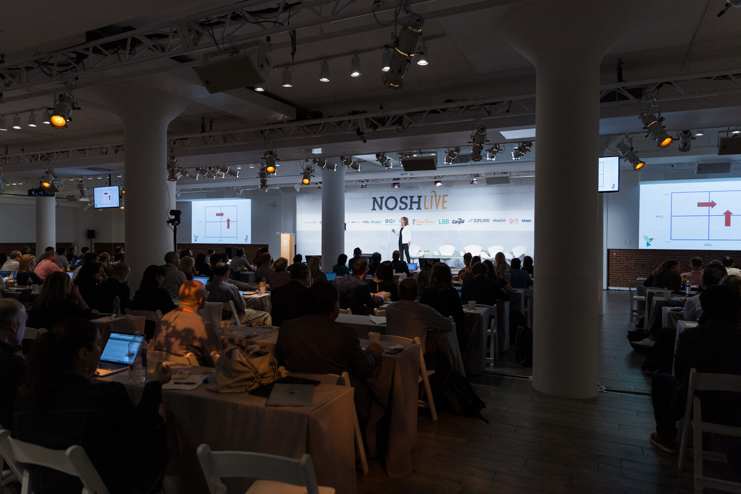 Wide view of the dark main hall with a speaker at the front and slides on the screens