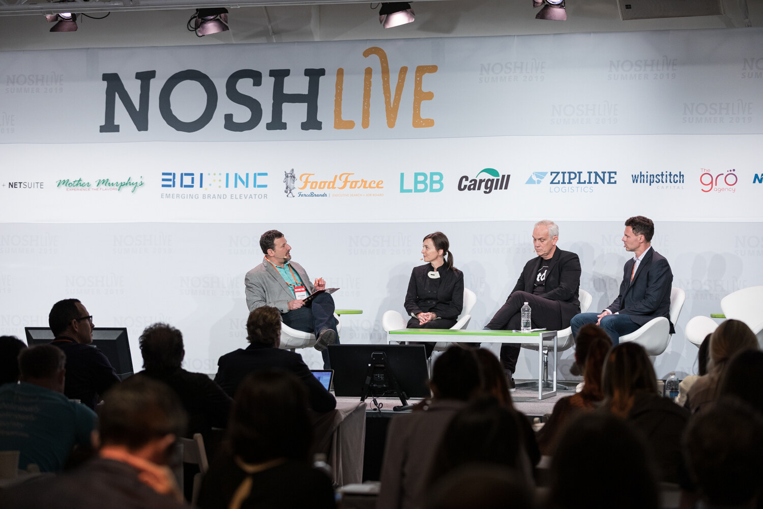 Wide view of the Brokers session on stage in front of the NOSHLIVE wall with the audience in the foreground