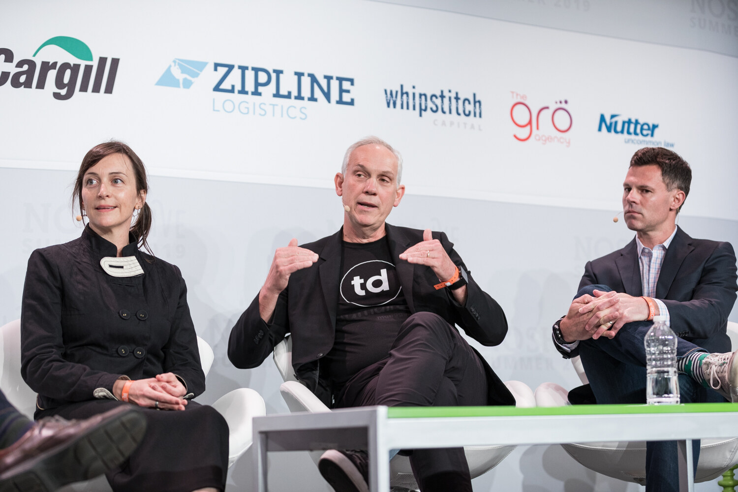 Man in a black td T-shirt gesturing with both hands while the woman in a black jacket and the man in a navy blazer listen