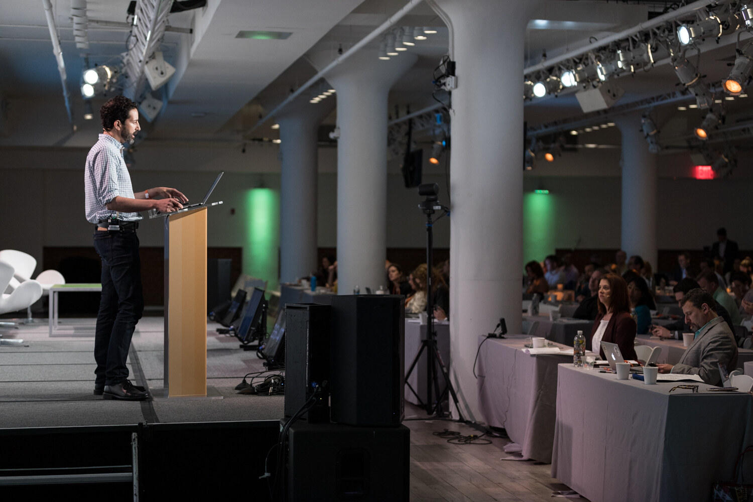 Bearded man at the lectern seen across the hall with columns, green lights and draped attendee tables