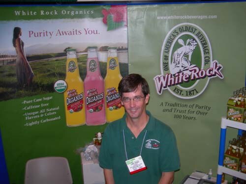 A man with glasses and a green shirt stands at a booth with White Rock Organics banners showing bottled sodas and the White Rock logo reading Est. 1871.