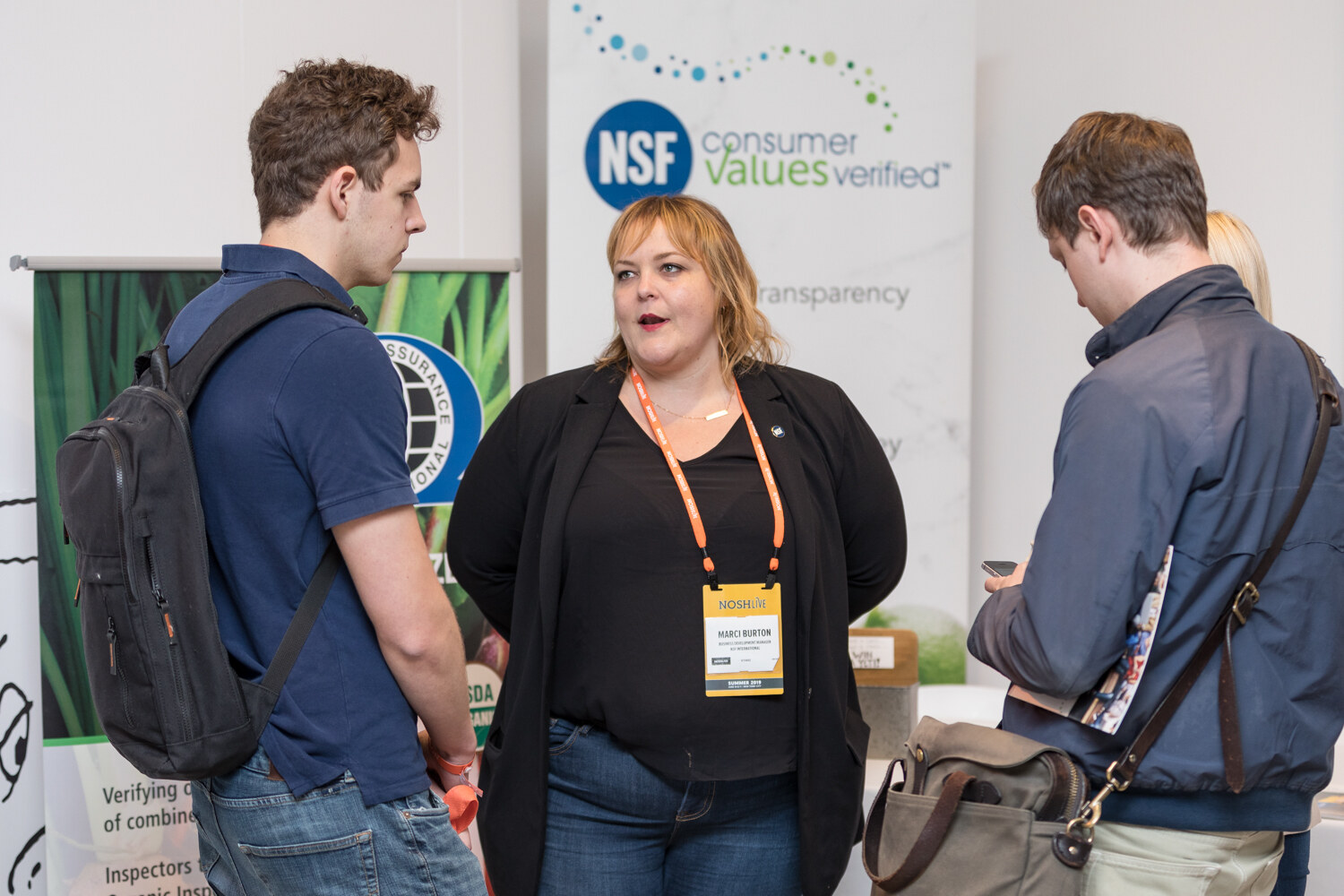 Woman in a black cardigan in front of an NSF consumer values verified banner talking with attendees