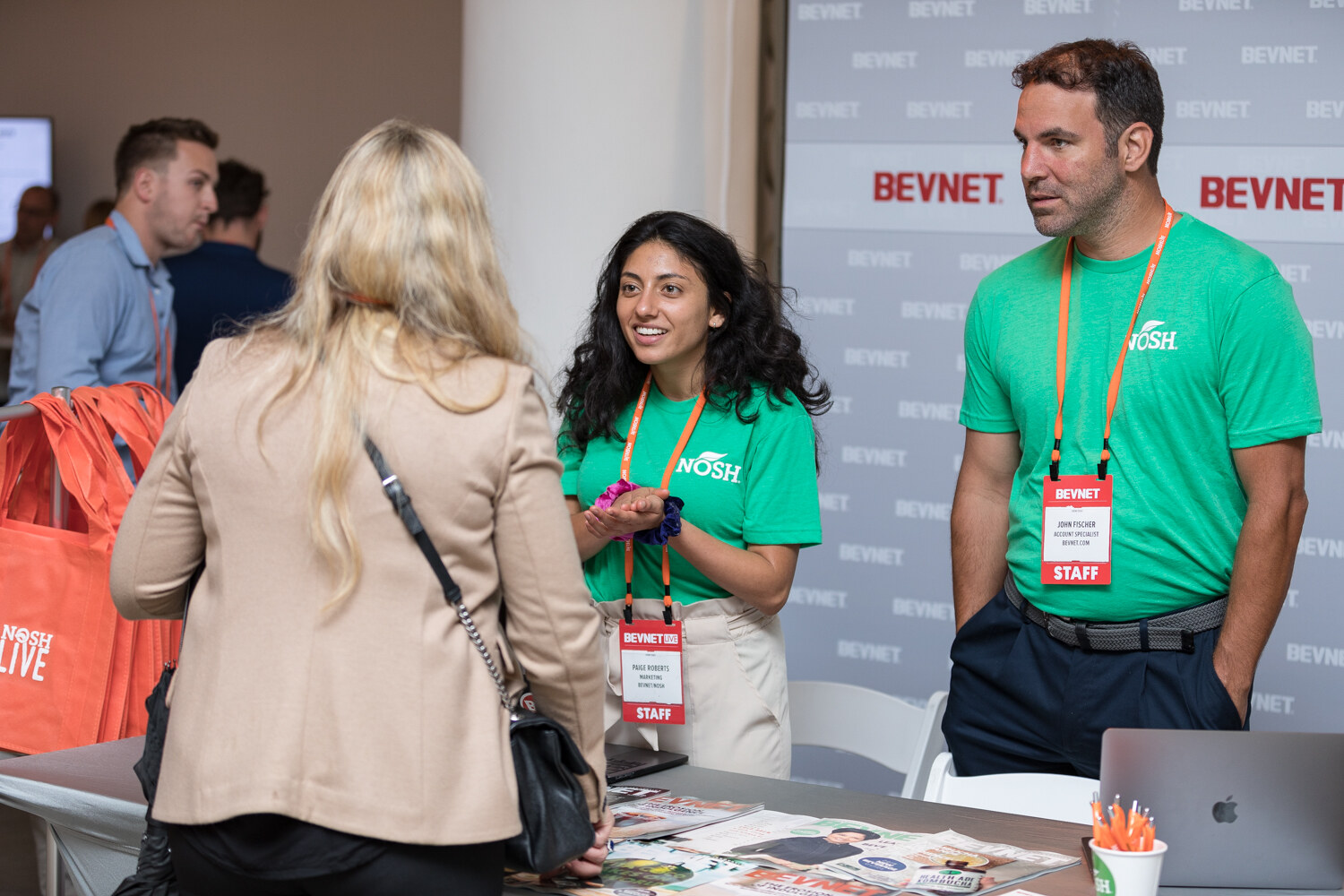 Two BEVNET staffers in green Nosh T-shirts at the media table in front of a BEVNET backdrop