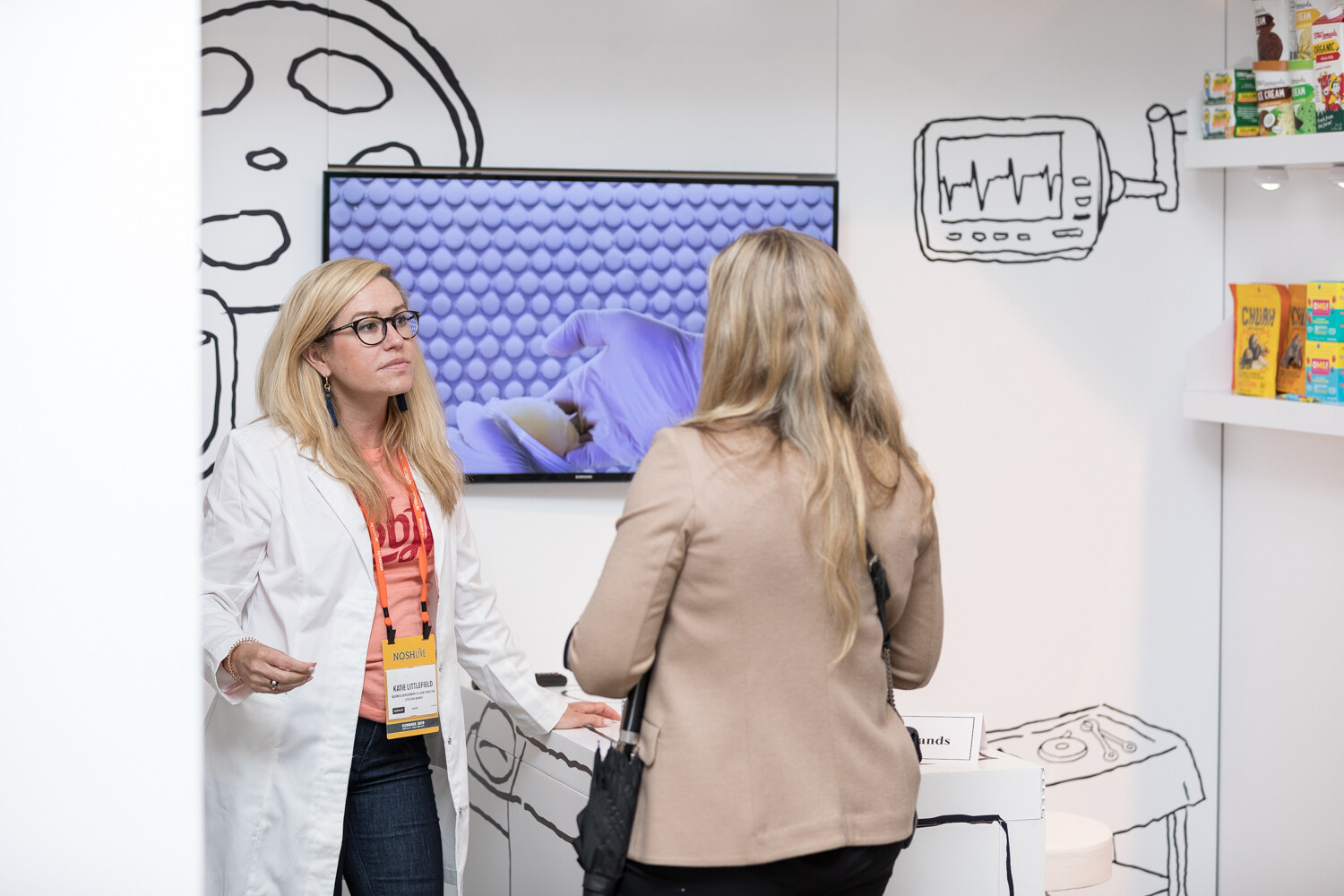 Staffer in a white lab coat at the Brand Surgery booth beside a woman in a tan jacket