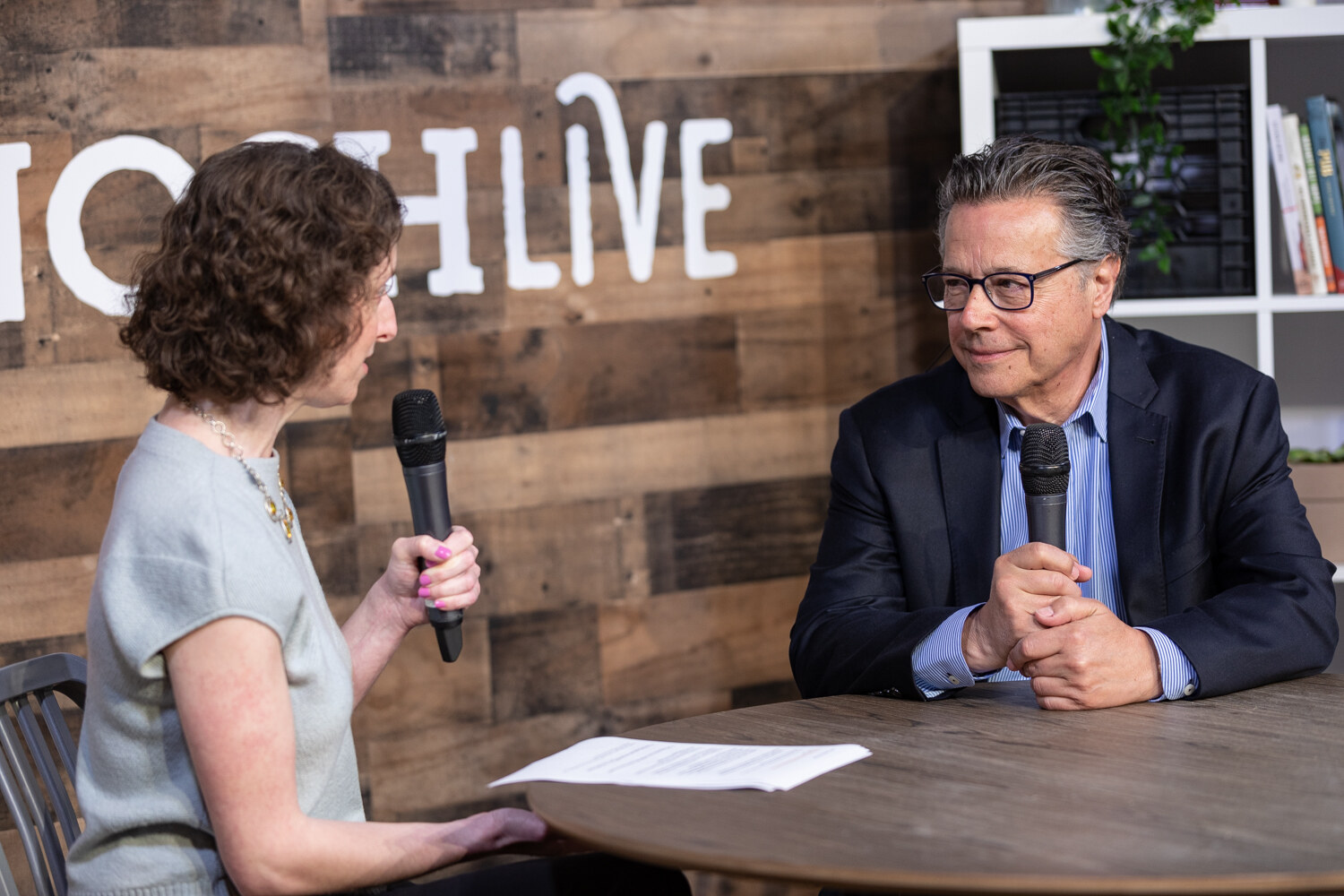 Curly-haired interviewer and the gray-haired man in glasses and a navy blazer at the round livestream table