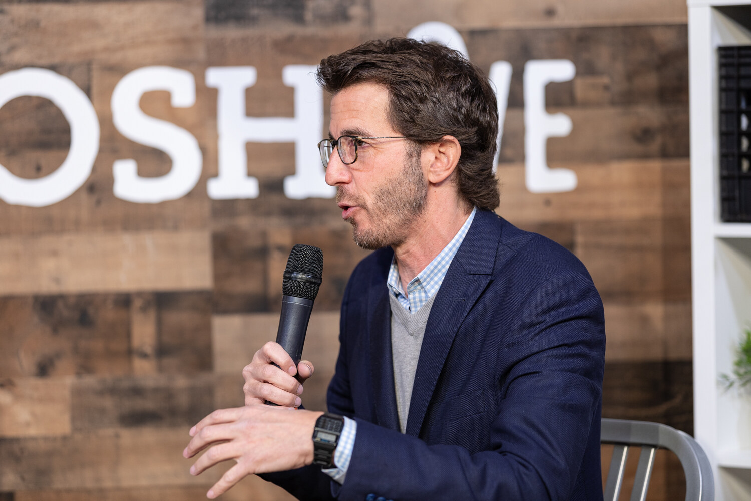 Man with wavy dark hair and glasses in a navy blazer and gray sweater speaking into a microphone