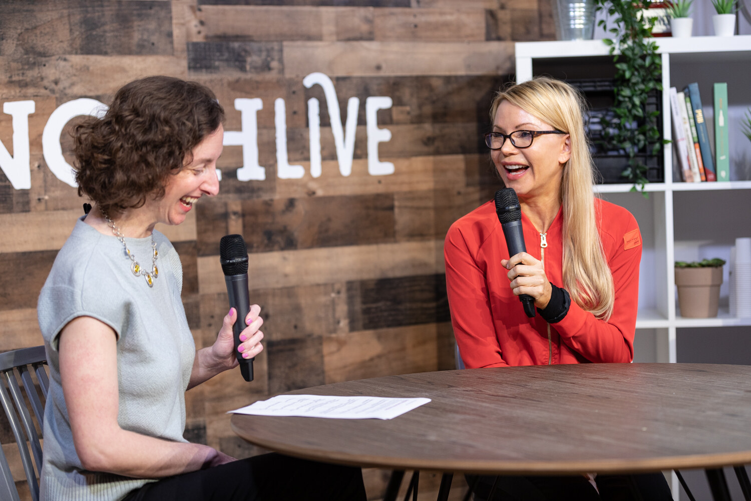 Curly-haired interviewer in a gray top laughing while the woman in a red zip jacket answers
