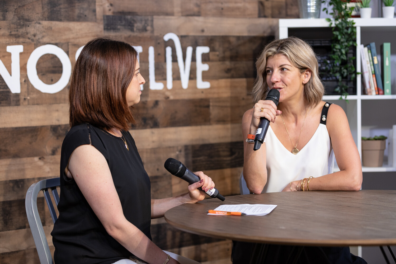 Interviewer in a black sleeveless top and a woman in a white camisole talking at the livestream table