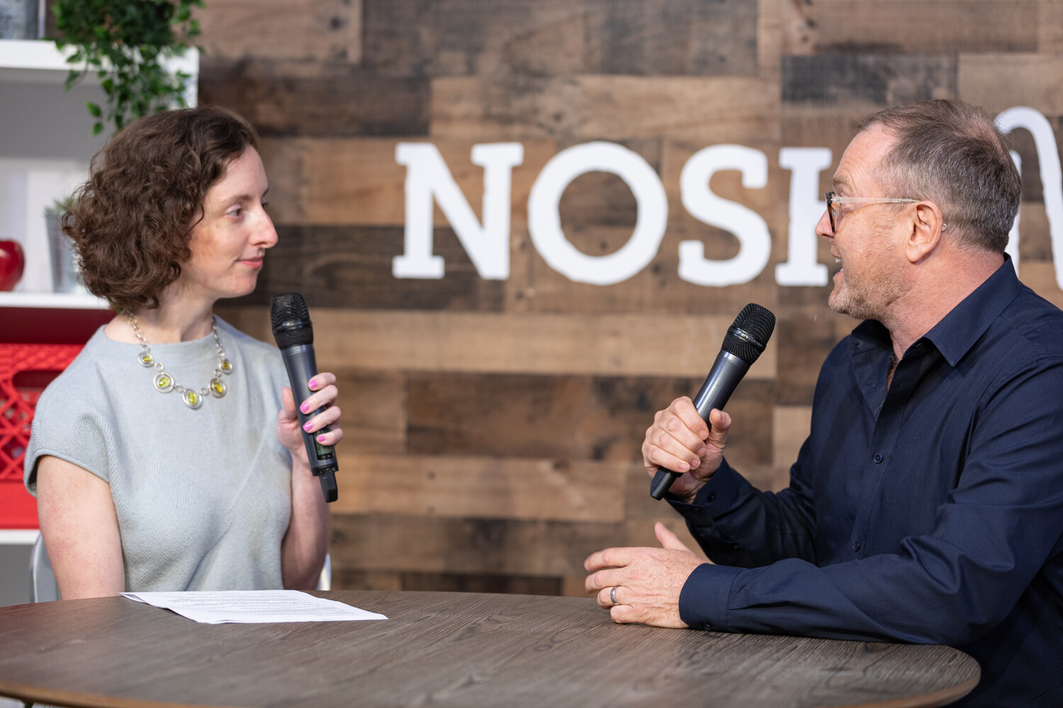 Curly-haired interviewer and a man with glasses in a navy shirt in conversation at the livestream table