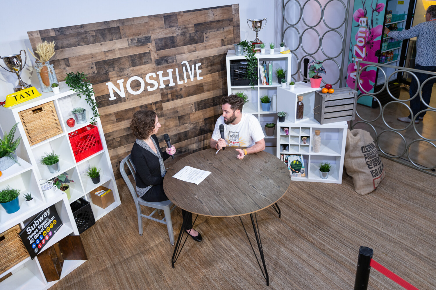Taste Radio livestream set seen from above, a curly-haired interviewer and a bearded guest in a graphic T-shirt at a round table in front of a NOSHLIVE wooden wall