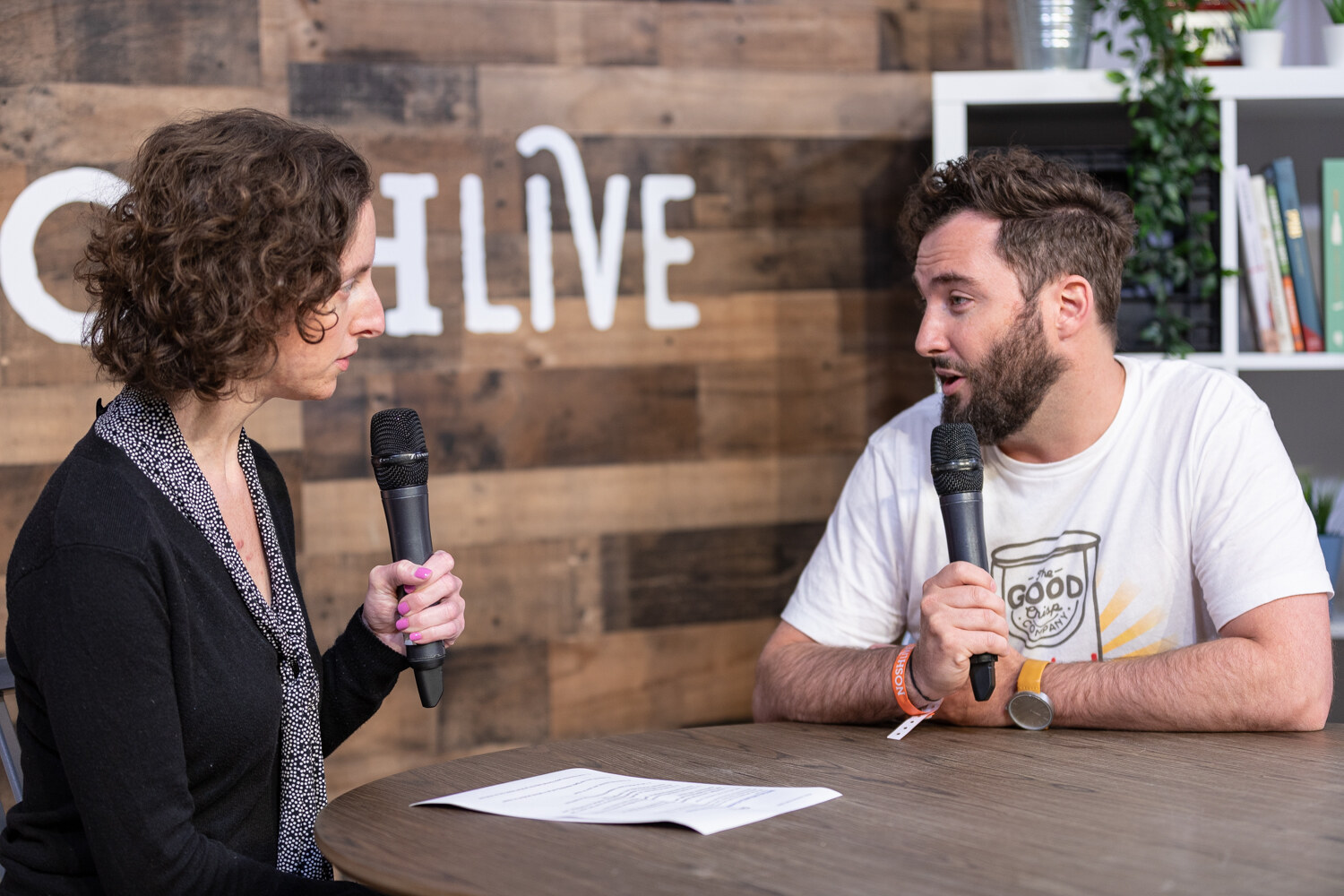 Curly-haired interviewer in a black cardigan and the bearded man in a Good T-shirt in conversation