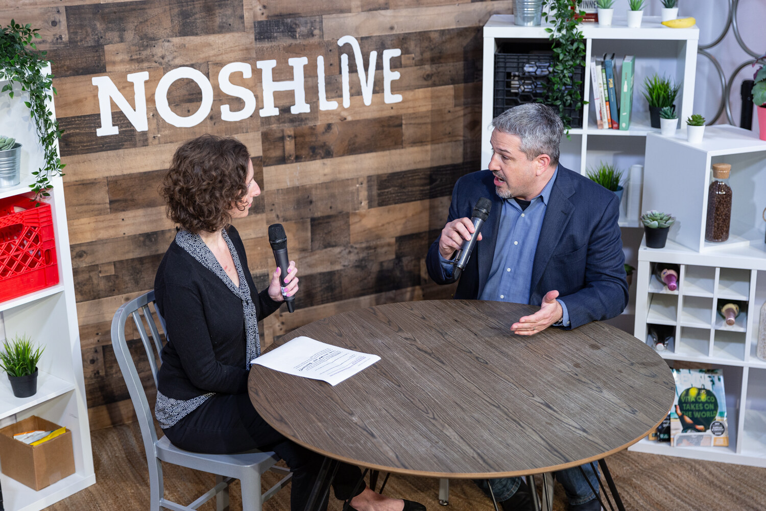 Curly-haired interviewer holding a microphone and a man in a navy blazer gesturing at the round table of the Taste Radio livestream set