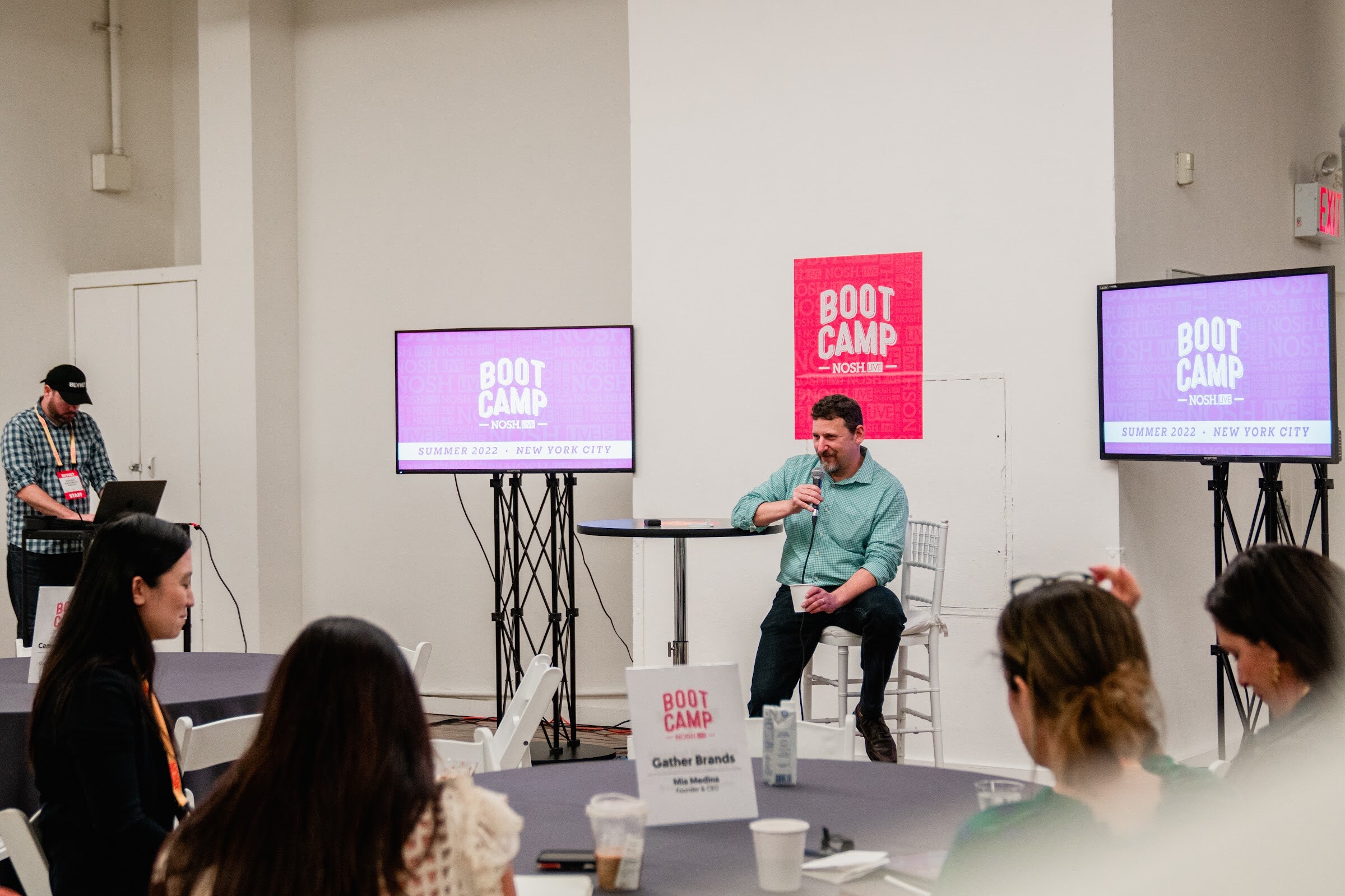 Man in a green shirt seated on a stool at the front of the room, two purple Boot Camp screens and attendees seated at tables.