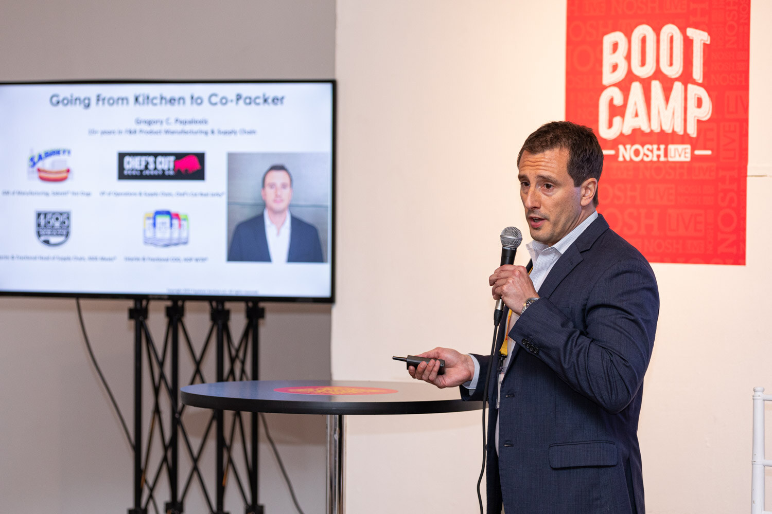 Man in a navy blazer holding a mic and clicker in front of a Boot Camp sign and a screen titled Going From Kitchen to Co-Packer.