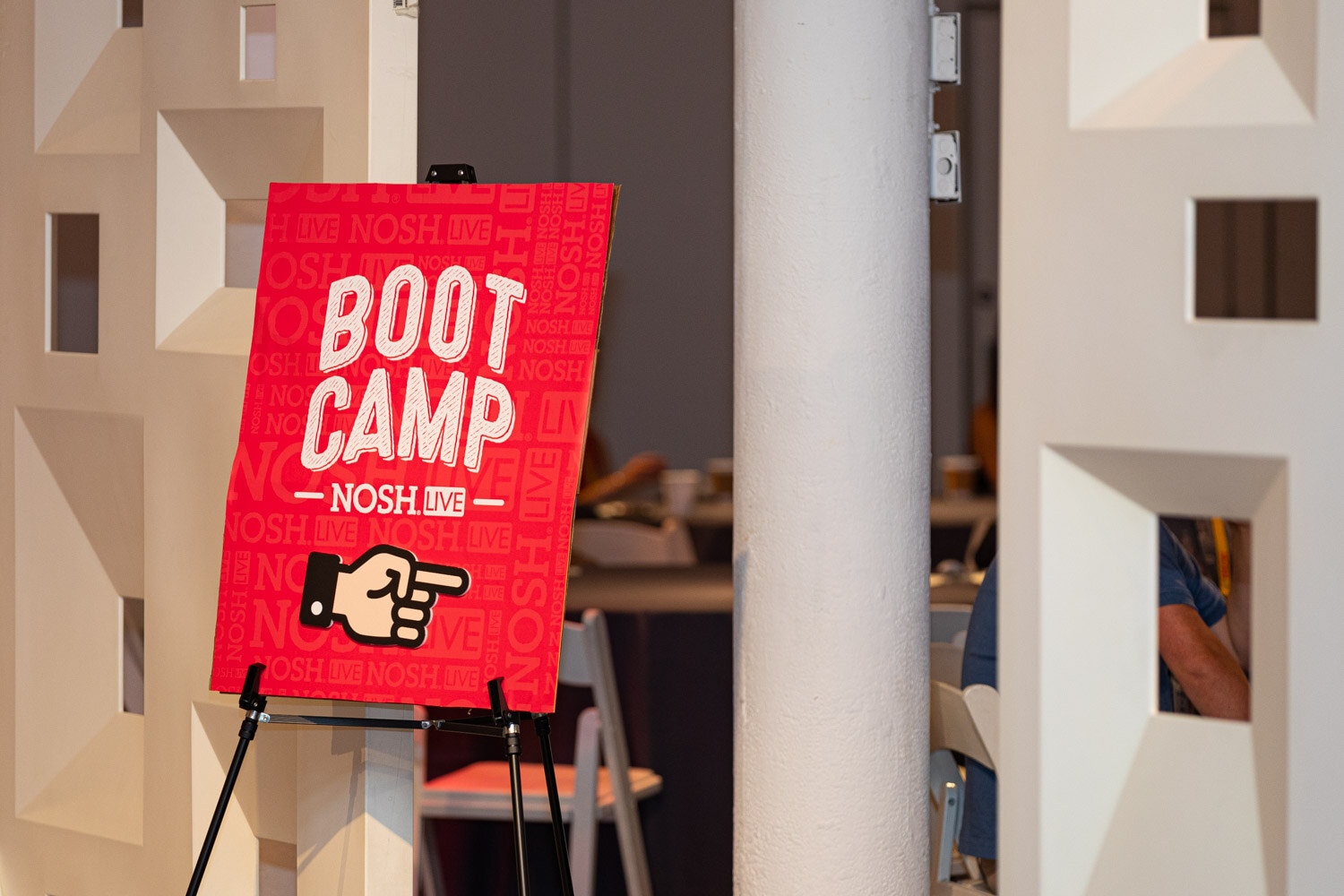 Red Nosh Live Boot Camp sign with a pointing hand on an easel beside a white column.