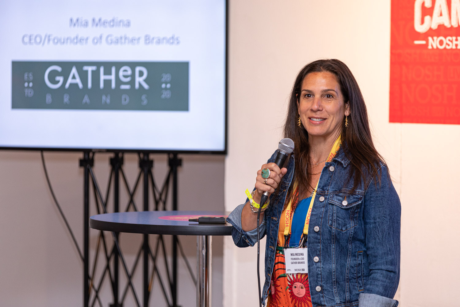 Woman in a denim jacket and bright dress with a mic beside a screen reading Gather Brands.