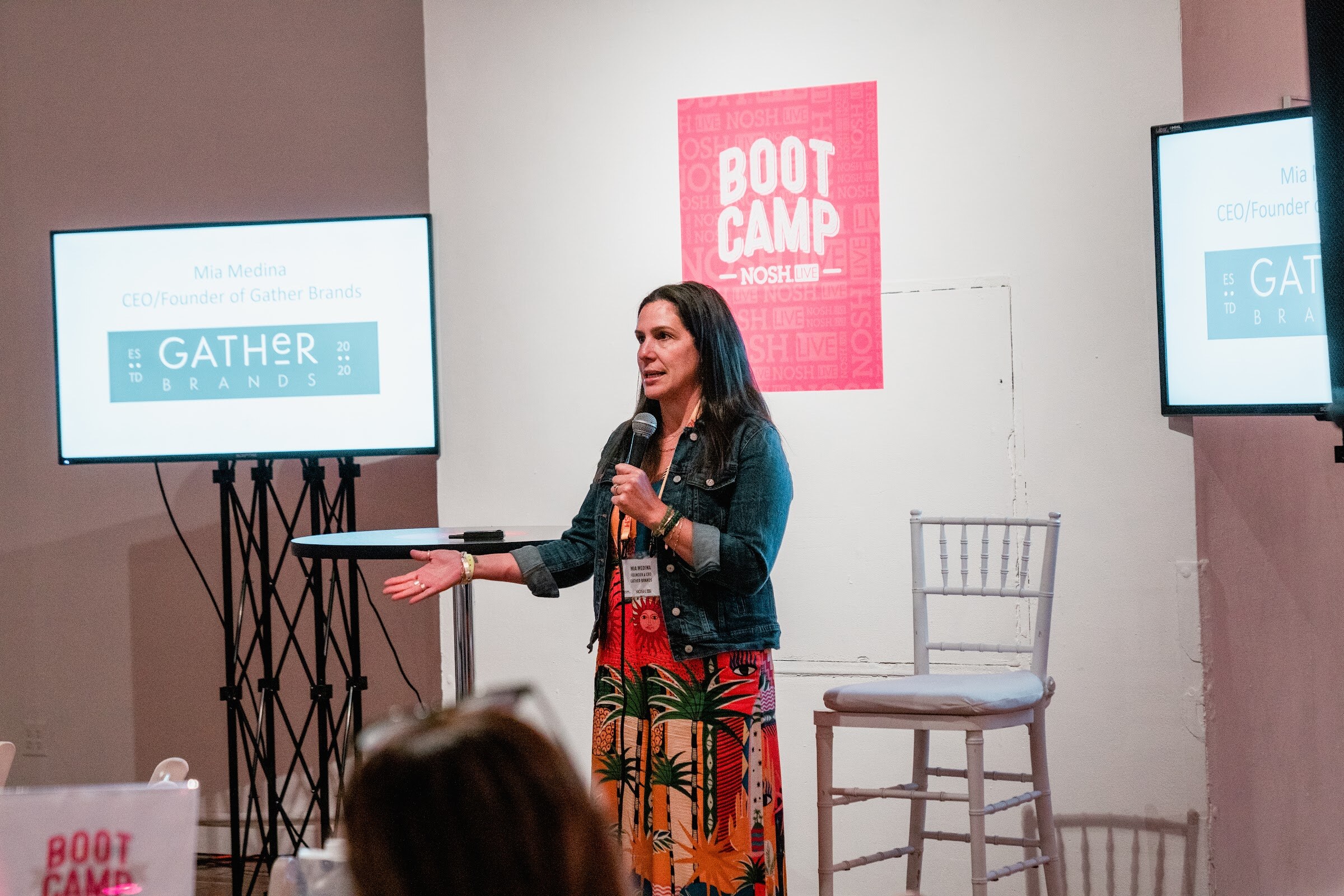 Woman in a denim jacket and tropical skirt with a mic, a screen reading Gather Brands and a Boot Camp sign behind her.