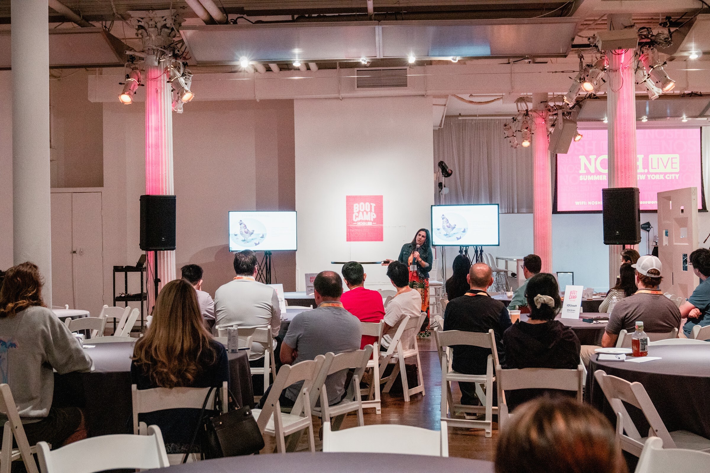 Wide room view with rounds of seated attendees, two screens, a red Boot Camp sign and pink-lit columns.
