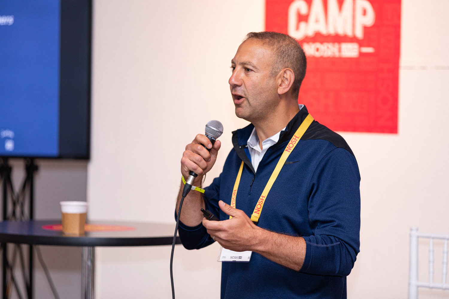 Man in a navy quarter-zip with a yellow lanyard holding a mic in front of a red Boot Camp sign.