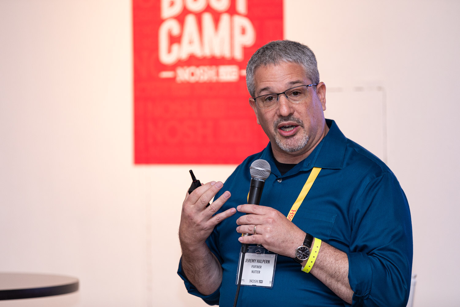 Man in a blue shirt with a lanyard holding a mic and clicker in front of a Boot Camp sign.
