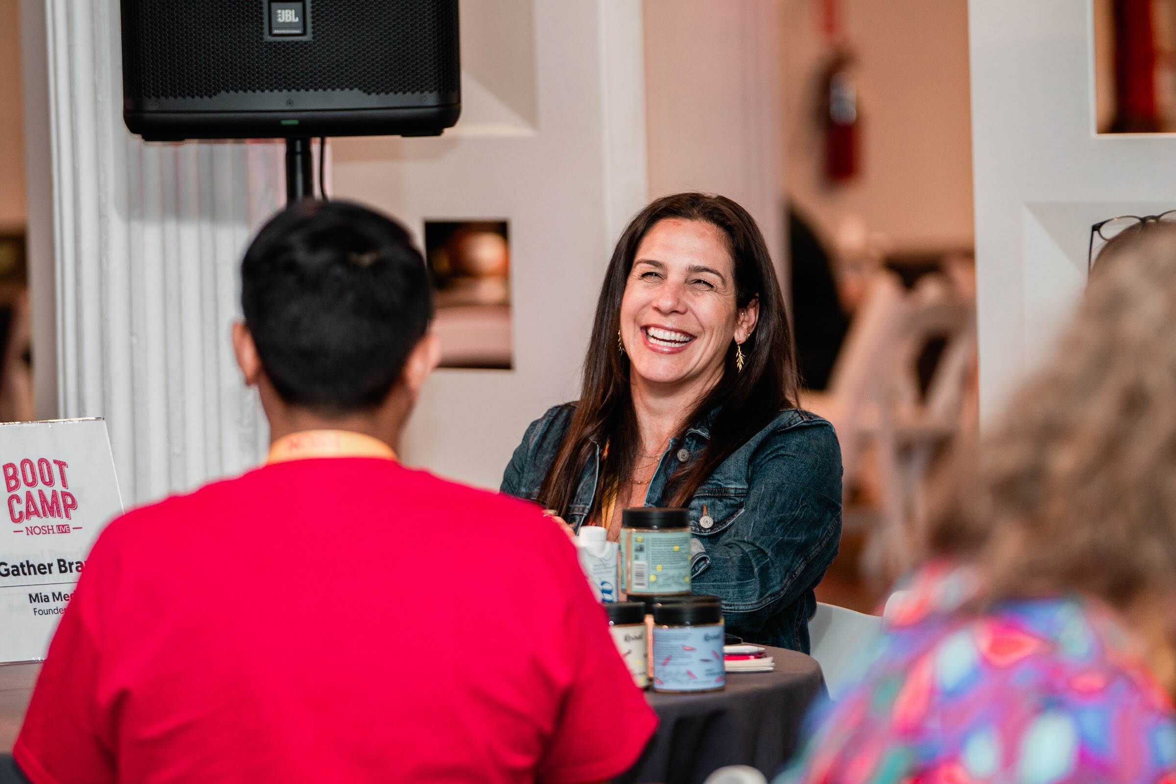 Woman in a denim jacket smiling at a table with jars of product, a man in a red shirt in the foreground, a Boot Camp sign at the left.