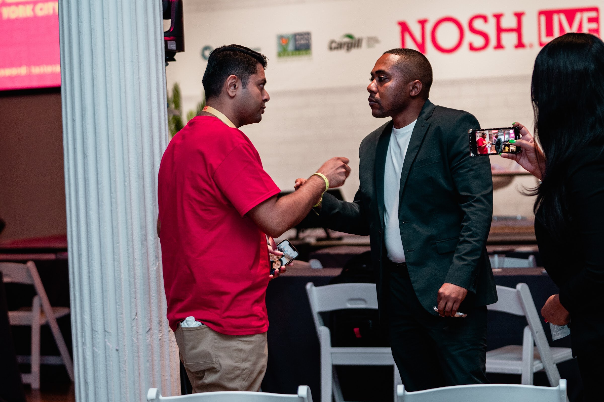 Man in a red shirt and man in a dark suit shaking hands in front of a white column and the Nosh Live wall.