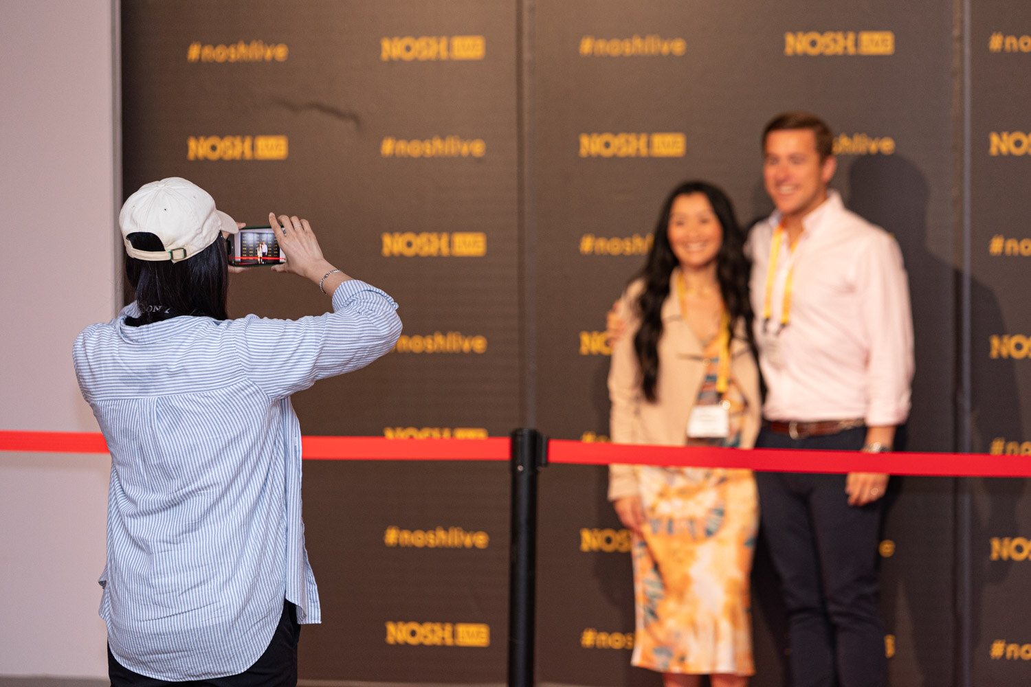 Woman in a floral dress and man in a pink shirt at the black #noshlive backdrop, a person in a white cap photographing them.