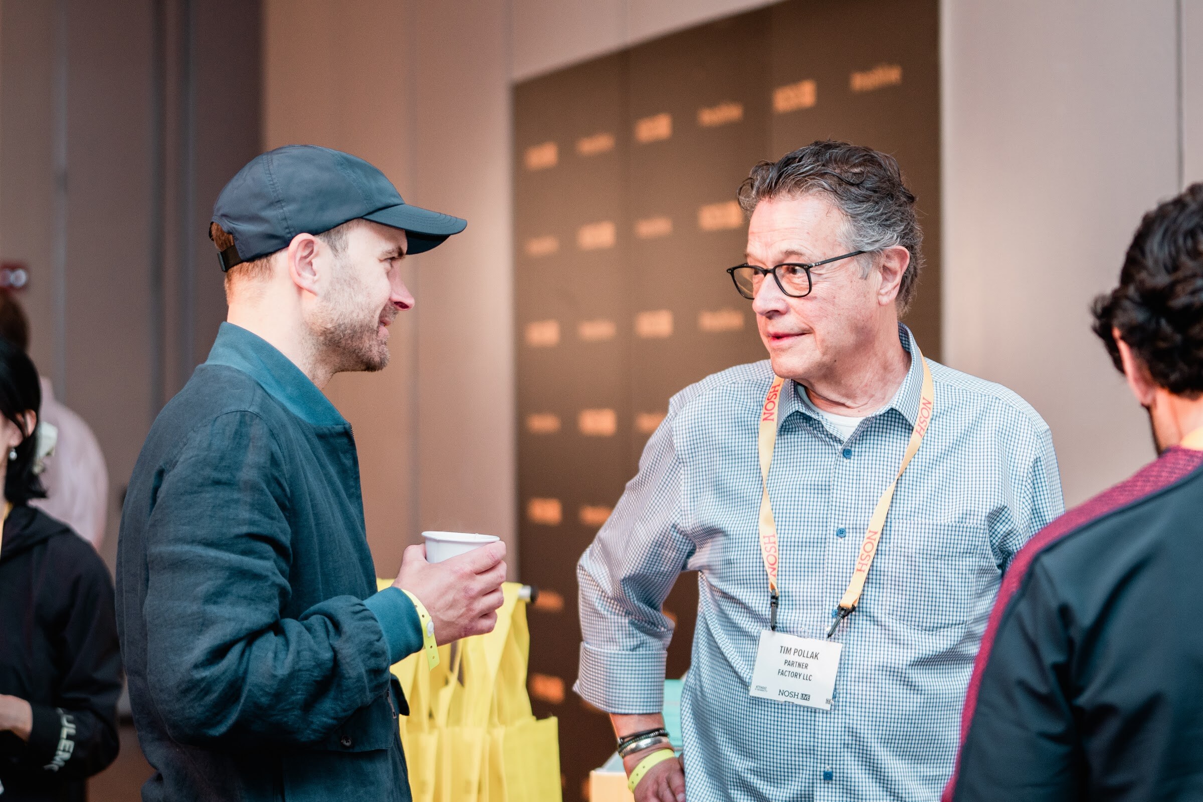 Man in a navy cap and teal jacket and man in glasses and a blue shirt with a lanyard talking, yellow tote bags beside them.