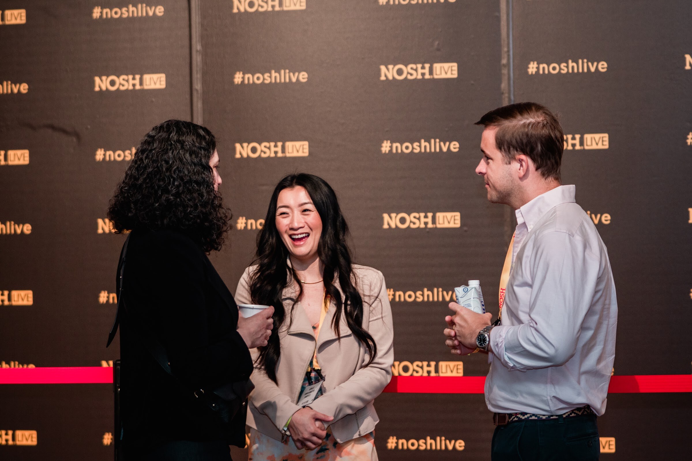 Woman with dark curly hair, a woman in a beige jacket and a man in a white shirt talking in front of the black #noshlive wall.