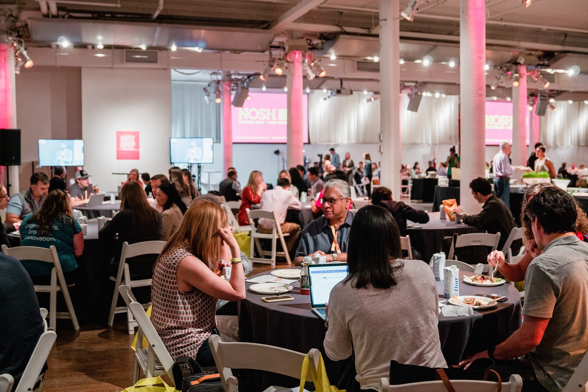 Wide view of the room full of round tables with attendees eating and pink screens at the back.