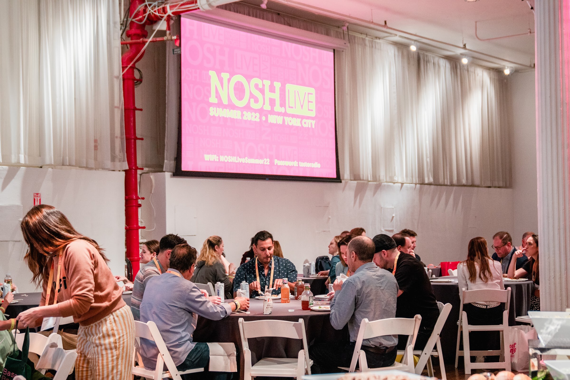 Crowded round tables in front of a large pink screen reading Nosh Live Summer 2022 New York City.