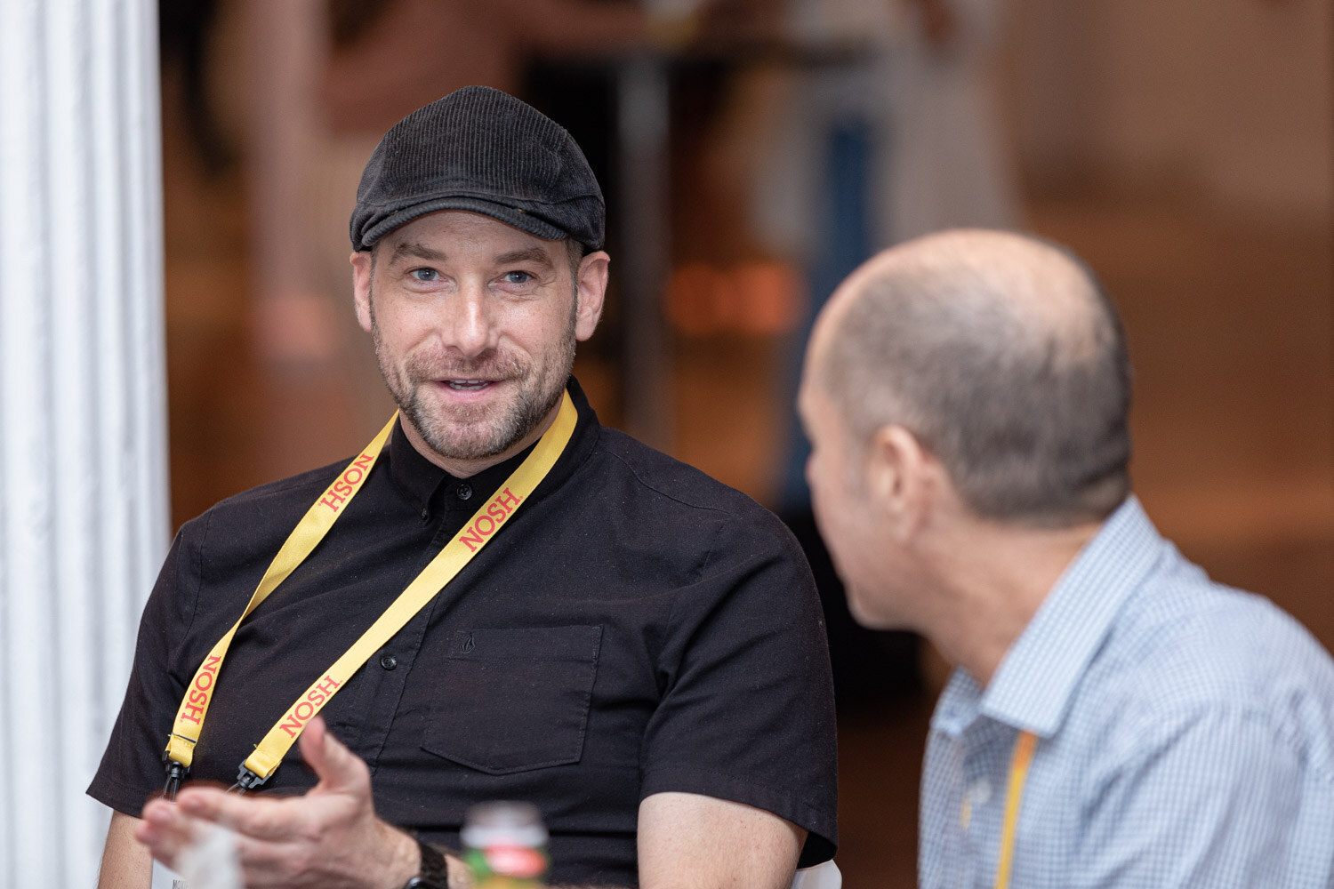 Smiling man in a black shirt and cap with a yellow lanyard talking with a man in a light blue shirt.