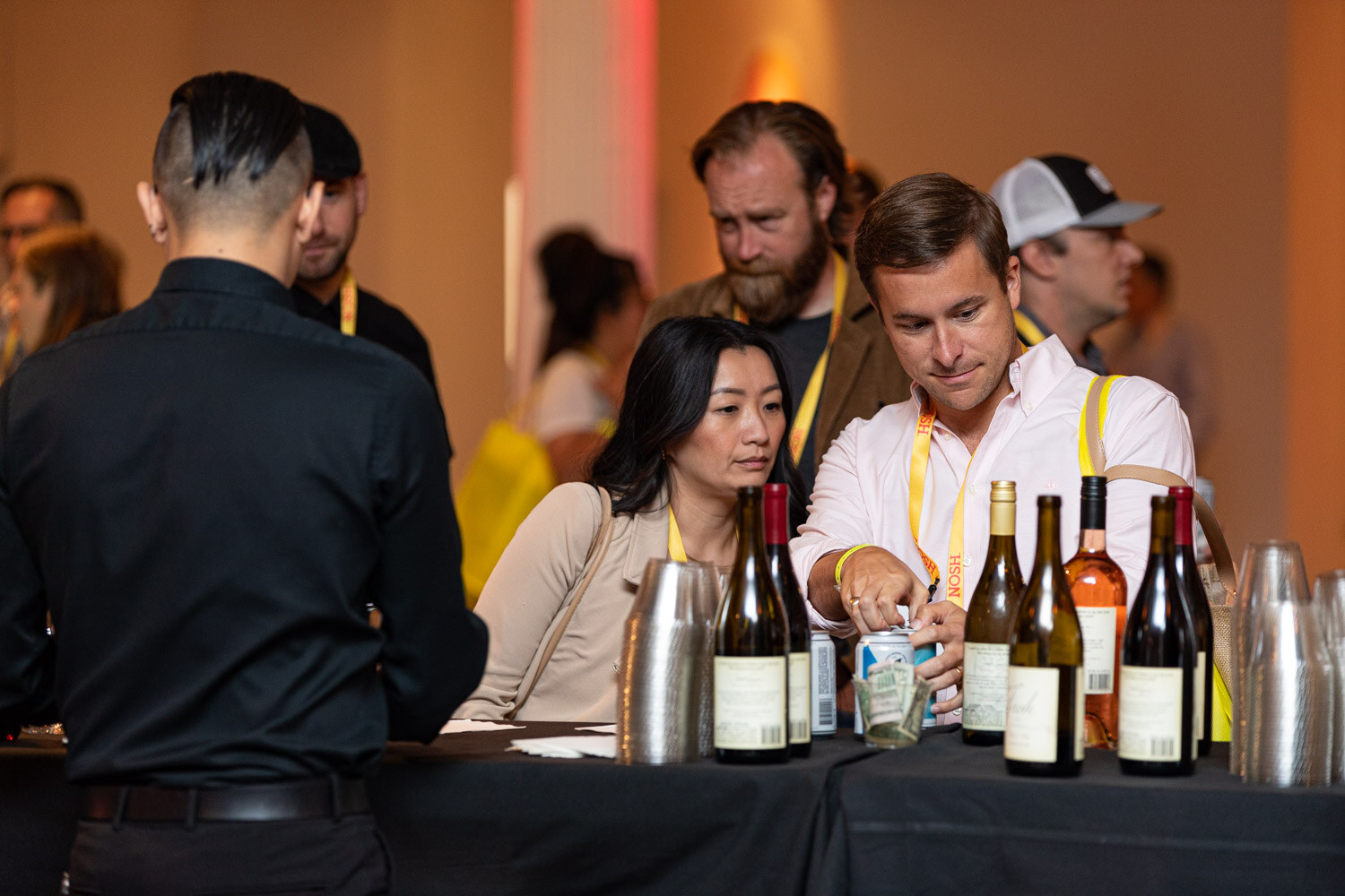 Black bar table with wine bottles and shakers, a bartender in black with his back to the camera and guests in front of him.