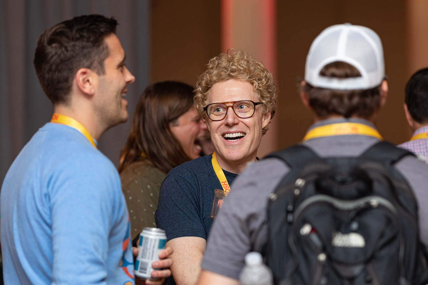 Man in a blue sweater and man with curly hair and glasses laughing, a man in a white cap and backpack in the foreground.
